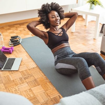 African american sportswoman doing abs on yoga mat near laptop in living room. African-American woman having an online training at home. Exercising on the floor